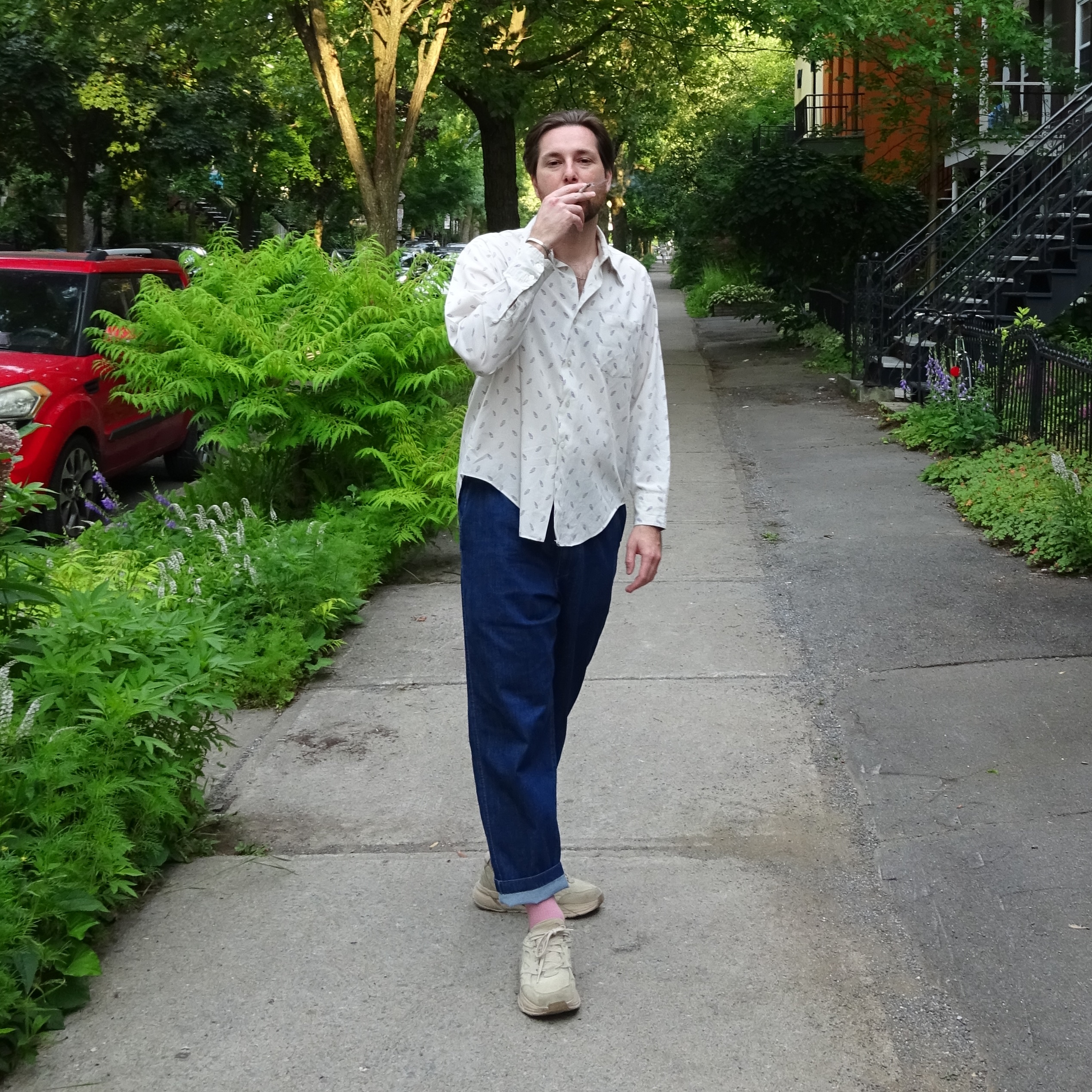 Image of elliot standing on a sidewalk in Montreal taking a puff of a joint with lots of green trees in the background